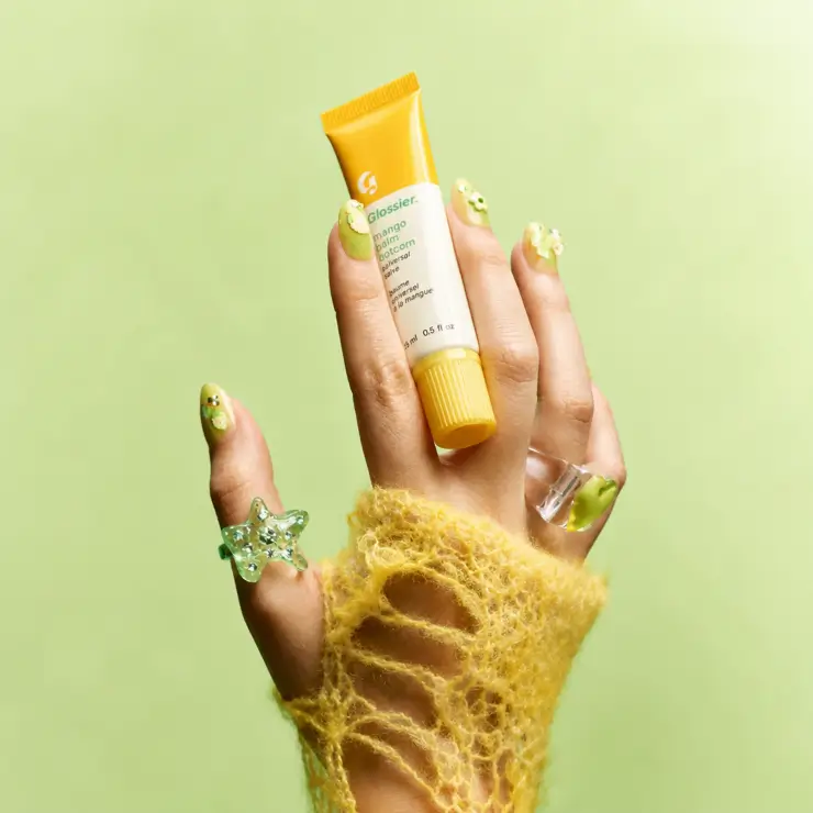 AI creative close-up beauty product photography of a hand wearing trendy rings and a yellow knit sleeve holding a yellow lip balm tube against a light green background